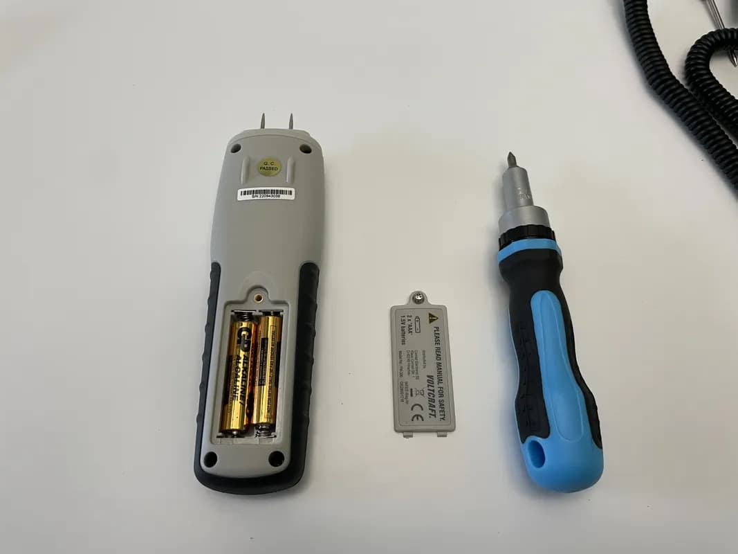Das Holzfeuchtemessgerät VOLTCRAFT FM-300 liegt mit aufgeschraubtem Batteriefach auf dem Tisch. Daneben liegen die Abdeckung und ein Schraubendreher.