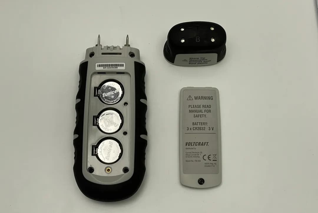Das Holzfeuchtemessgerät VOLTCRAFT FM-400 liegt mit aufgeschraubtem Batteriefach auf dem Tisch. Daneben liegt die Abdeckung.
