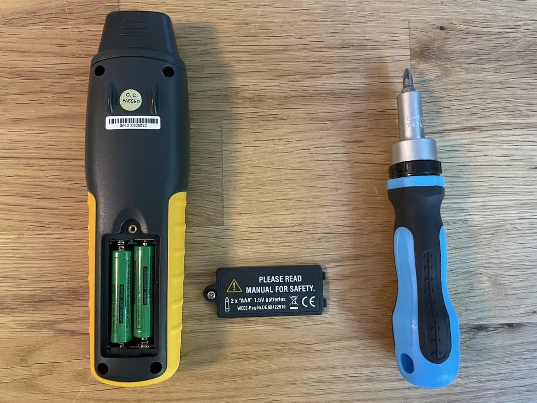 Das Holzfeuchtemessgerät TFA Dostmann HumidCheck PRO liegt mit aufgeschraubtem Batteriefach auf dem Tisch. Daneben liegen die Abdeckung und ein Schraubendreher.