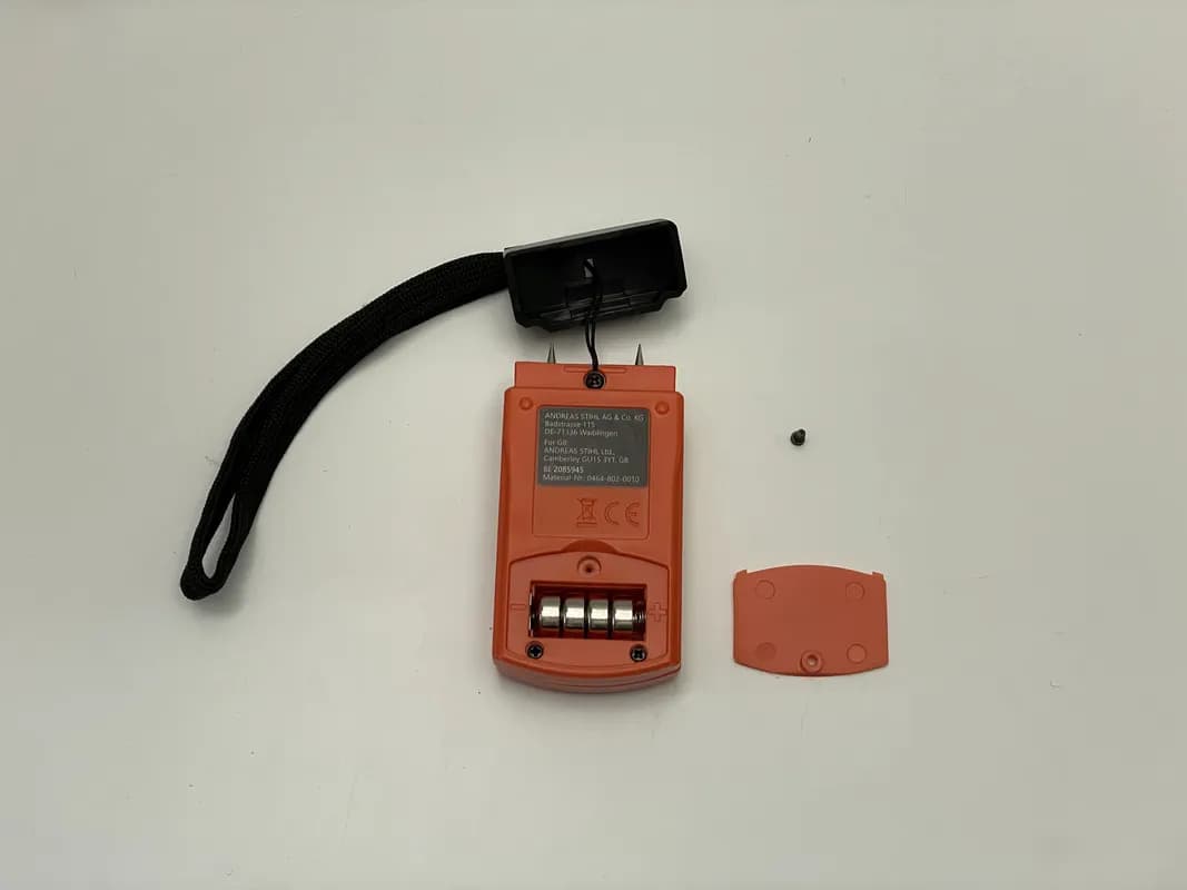 Das Holzfeuchtemessgerät von Stihl liegt mit aufgeschraubtem Batteriefach auf dem Tisch. Daneben liegt die Abdeckung.
